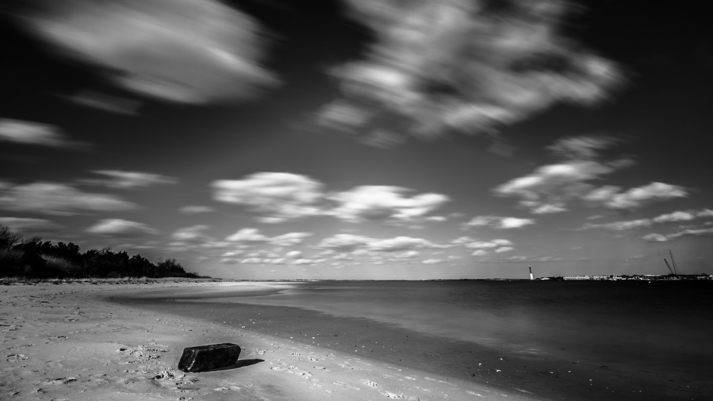 An afternoon long exposure photograph taken from High Bar Harbor in Barnegat Light overlooking east toward the Barnegat Lighthouse. In this empty bay beach scene, the low level clouds streak across this black and white photograph from left to right.