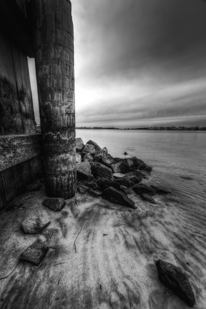 A seven frame black and white composite exposure of a wooden bulkhead and mounded jetty rock define the portrait orientation scene with Barnegat Bay expanding off to the right.