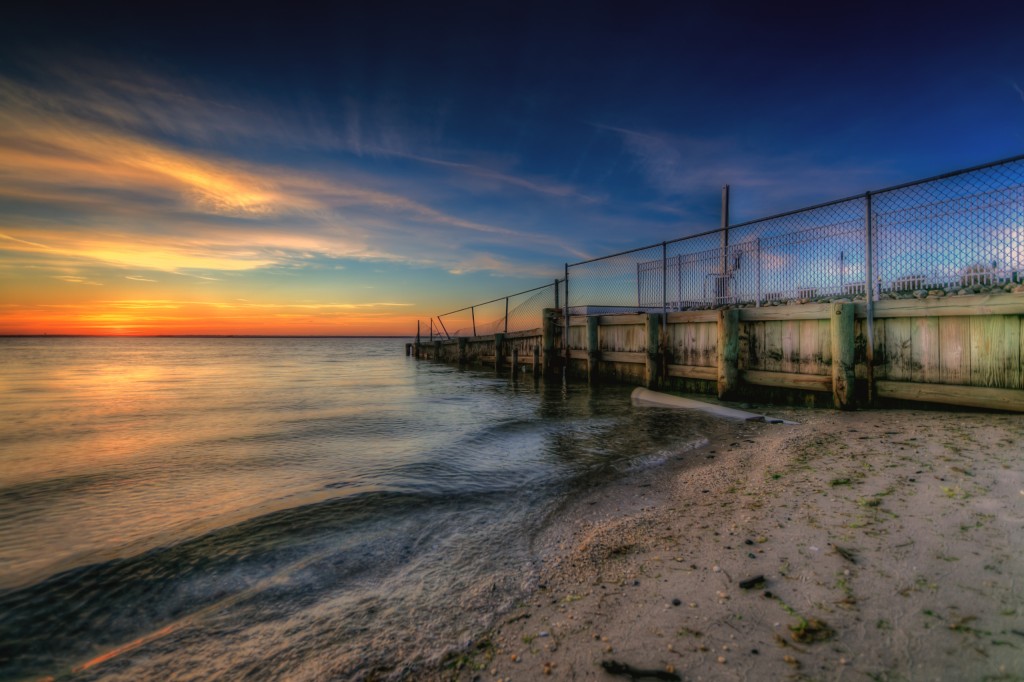 HDR photograph taken just before sunset at Sunset Park in Surf City, New Jersey. This photograph features a bulkhead capped by fence and fiery sunset colors over Barnegat Bay.
