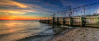 HDR photograph taken just before sunset at Sunset Park in Surf City, New Jersey. This photograph features a bulkhead capped by fence and fiery sunset colors over Barnegat Bay.