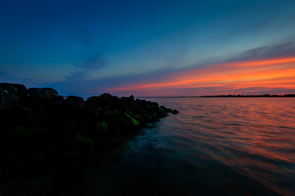 An HDR photograph taken at sunset from along the jetty at Sunset Point in Ship Bottom, New Jersey. The in water perspective hugs the darkened jetty rock and frames the colored sky and water.