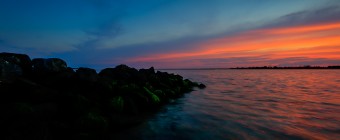 An HDR photograph taken at sunset from along the jetty at Sunset Point in Ship Bottom, New Jersey. The in water perspective hugs the darkened jetty rock and frames the colored sky and water.