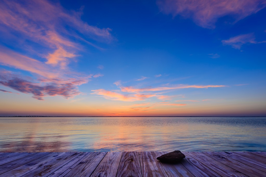 A late evening HDR photograph taken just after sunset from Sunset Park in Surf City, New Jersey. The exposure looks out over a very calm Barnegat Bay and features the unfinished wood of the dock at the edge of the park with a small stone in the foreground.