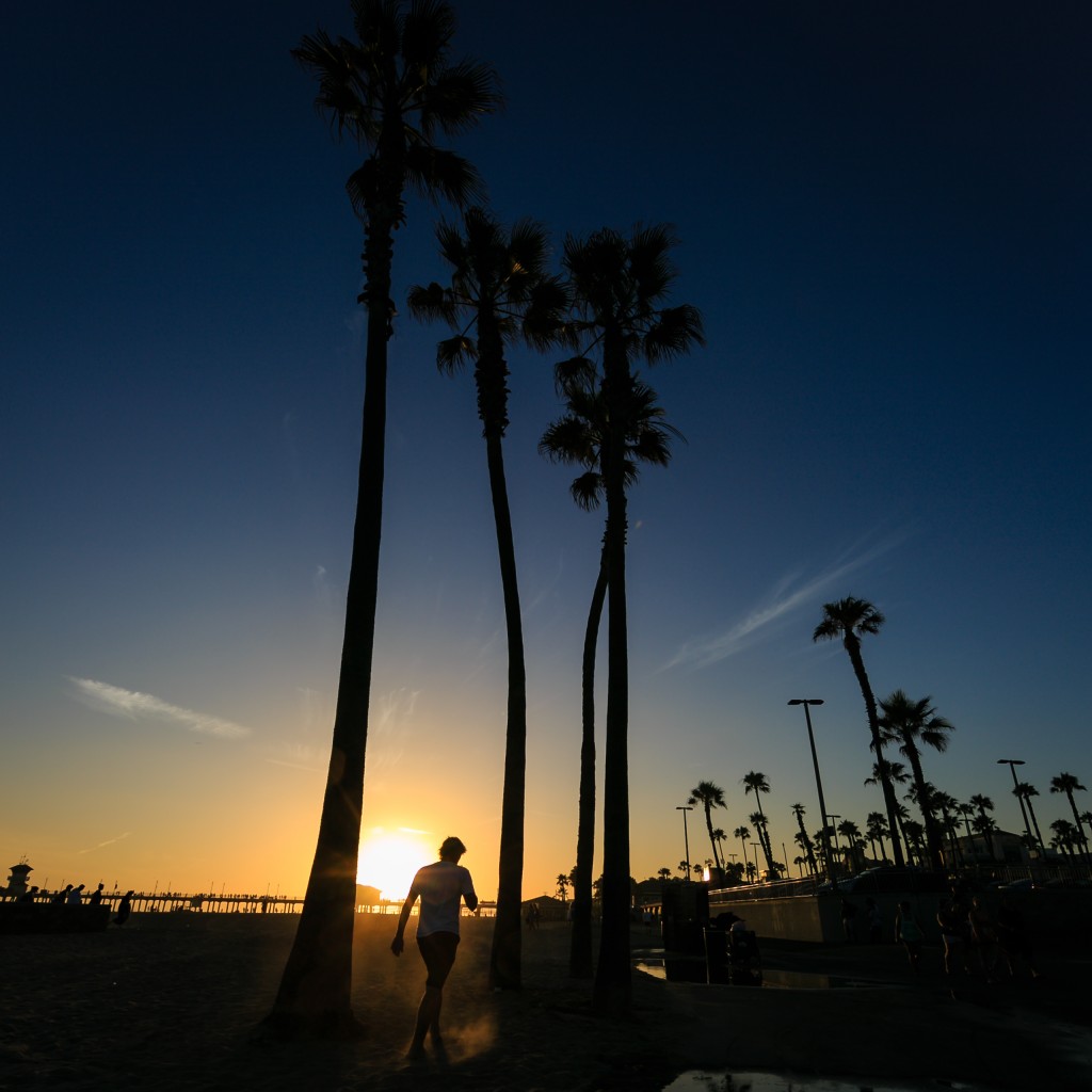 A sunset photograph taken during peak summer at the Huntington Beach, California, pier. A young man kicks up dust as he walks between palm trees with beautiful twilight approaching. 