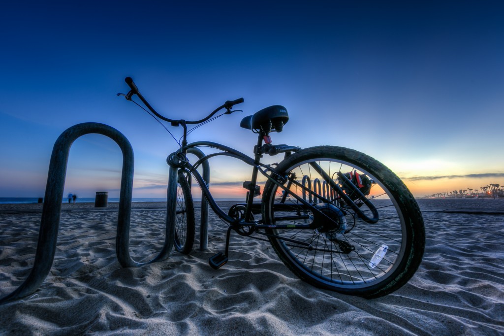 An HDR photograph taken just after sunset during blue hour along the shores of Huntington Beach California. The foreground is marked by a sweet beach cruiser bicycle locked to a beach bound bike rack. The Pacific Coast Highway lies away in the distance.