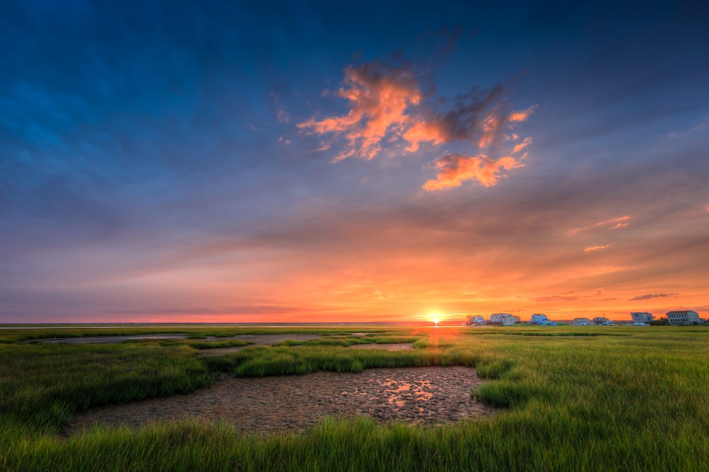 A subtle lens flare sends a rich  golden glow across the lush green salt marsh in this stunning HDR sunset photograph.