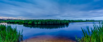 An evening HDR photograph of a winding waterway adjacent to Dock Road in West Creek, NJ. The blue tone picture features a small beach opening between long marsh grasses with a looming bank across the calm water.