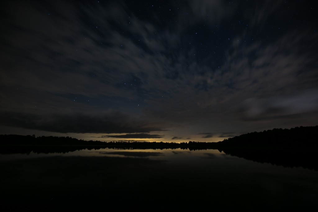 A long exposure wide angle landscape photograph taken at night just in front of the lake at Stafford Forge Wildlife Management Area. Low clouds race across a sky that's back-dropped with stars.