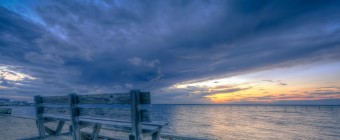 A cross processed wide angle HDR landscape photograph of a park bench, beach sand, and Barnegat Bay just at sunset. Dramatic clouds dominate the sky and a cool blue tone brings a real calming influence to the image.