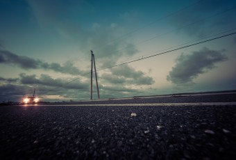 A cross processed street level photograph of a lone car approaching, headlights on. Power lines and clouds mark the background
