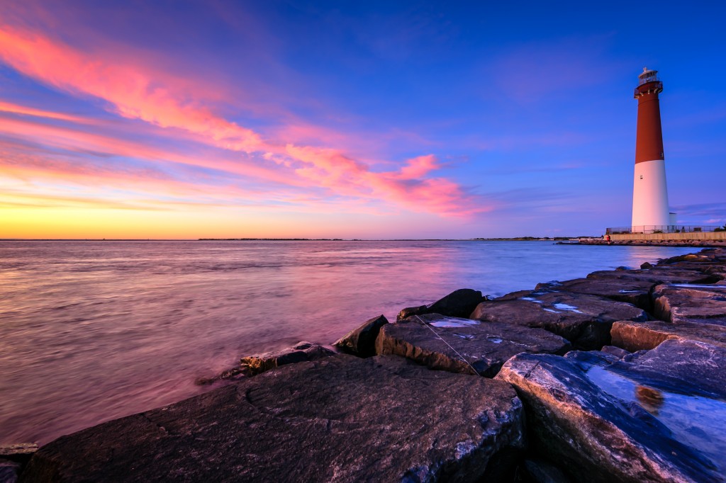 An HDR photograph of Barnegat Lighthouse taken from the jetty rock at blue hour overlooking majestic pastel clouds over Barnegat Bay. 