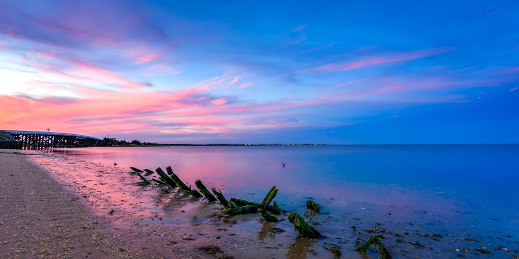 A serene coastline photographed at blue hour and processed for HDR; algae laden driftwood pieces mark the foreground with wisps of pink clouds setting beyond an old bridge and cool blue skies.