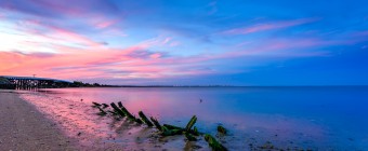 A serene coastline photographed at blue hour and processed for HDR; algae laden driftwood pieces mark the foreground with wisps of pink clouds setting beyond an old bridge and cool blue skies.
