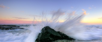 A blue hour photograph featuring ocean spray bursting behind a lone foreground jetty rock. sending water in all directions
