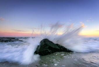 A blue hour photograph featuring ocean spray bursting behind a lone foreground jetty rock. sending water in all directions