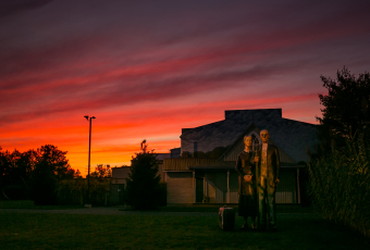 A photograph of a fiery sunset backlighting the American Gothic display at New Jersey's Grounds for Sculpture. True to the look of the original painting, here the famous farmers are depicted in larger than life cast bronze statuary.