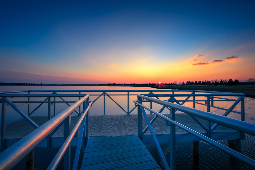 An HDR sunset photograph taken from the aluminum dock overlooking Bass River in New Gretna, New Jersey. With the metal dock marking the foreground, a rich pastel glow colors the sky and water on this near cloudless evening. 