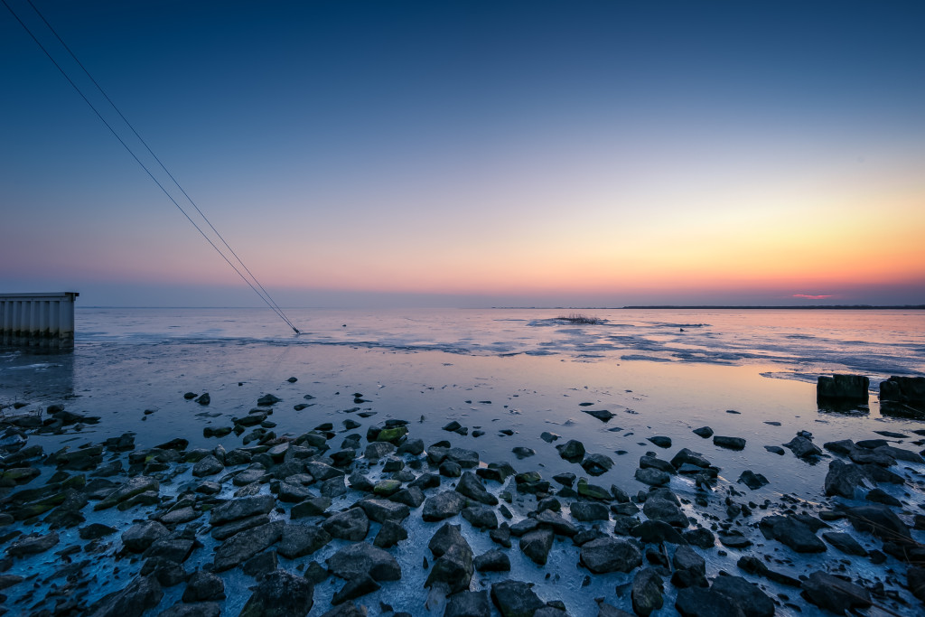 An HDR blue hour photograph of a stony bayshore mixed with thin sea ice in the foreground and angle guy-wires in the mid ground plunging into the frozen bay. 