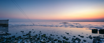 An HDR blue hour photograph of a stony bayshore mixed with thin sea ice in the foreground and angle guy-wires in the mid ground plunging into the frozen bay.