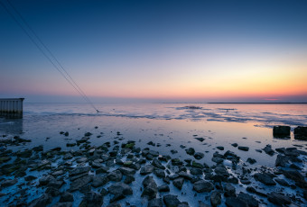 An HDR blue hour photograph of a stony bayshore mixed with thin sea ice in the foreground and angle guy-wires in the mid ground plunging into the frozen bay.
