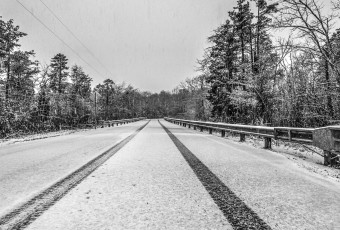 Light snow falls on an empty roadway marked only by a lone set of tire tracks. Flanking the road are guardrails and pine trees to either side of this black and white photograph.