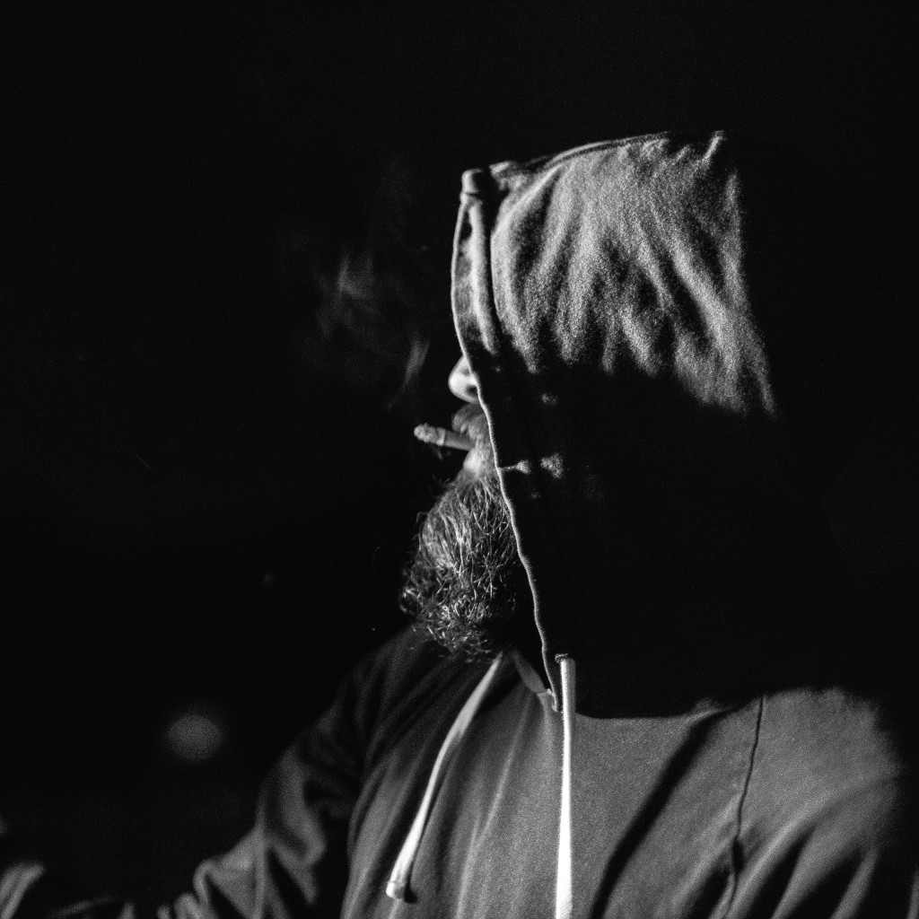 Ceramicist and potter, a hooded Jeff Ruemeli smokes the last of his cigarette as he stokes the soda kiln fires in this low key black and white portrait.