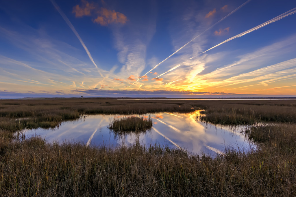 Contrails line the sky over a reflective marsh tide pool at sunset