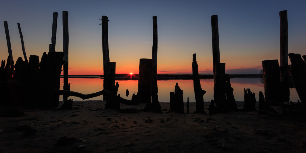 Wide angle landscape photograph of still water and silhouette dock remains at sunset