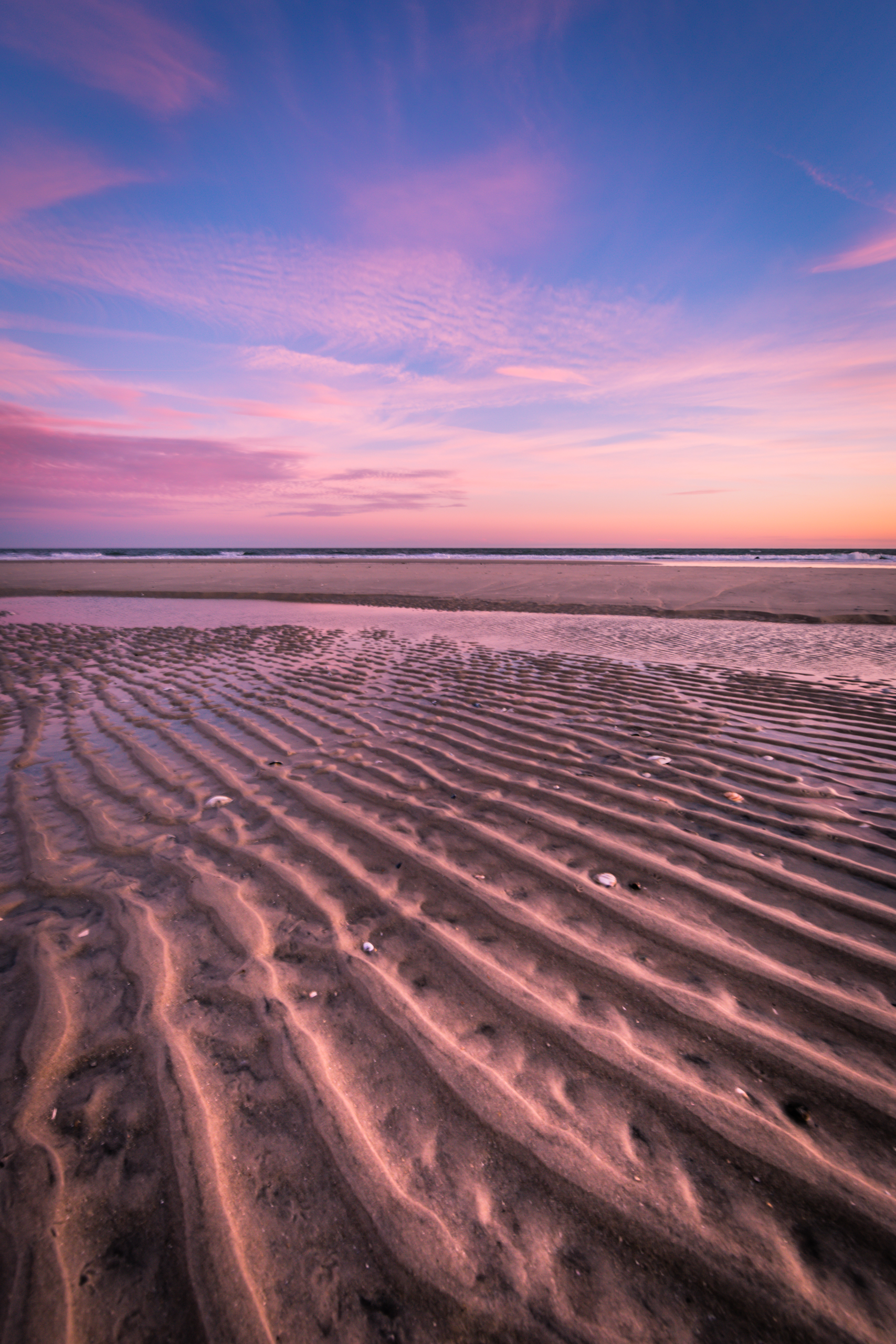 Vertical orientation photograph with strong leading lines in the sand underneath a pastel sunset