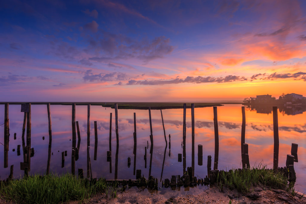 Sunset photo of layered pastel clouds, wood pilings, and a smooth water reflection