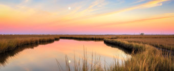 Sunset photo of a reflective moonrise over marsh amid pastel clouds.