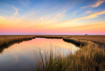 Sunset photo of a reflective moonrise over marsh amid pastel clouds.