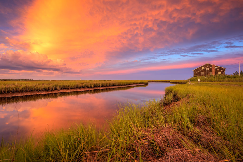  Explosive sunset photo over salt marsh, water, and house.