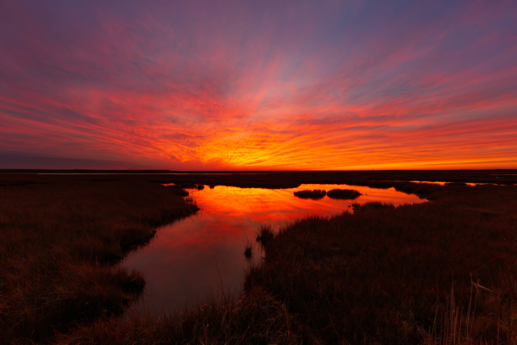 14mm photo of a burning red sunset smoldering over the winter salt marsh with sky reflections in the water. 