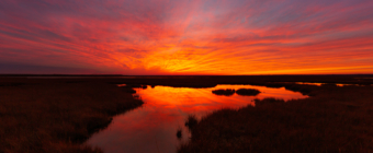14mm photo of a burning red sunset smoldering over the winter salt marsh with sky reflections in the water.