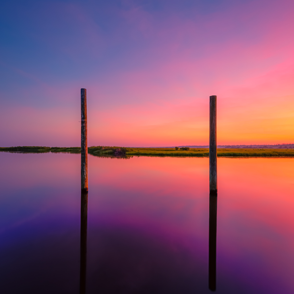 14mm square format photo of a potent pastel sunset reflecting over a glassy Cedar Run creek. Two vertical pilings mark the mid ground. 