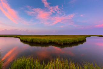 14mm wide angle photograph of an oxbow feature winding through the salt marsh. A pastel sunset sparkles in the sky, marsh grasses frame the foreground with clouds mirror reflected in the water.