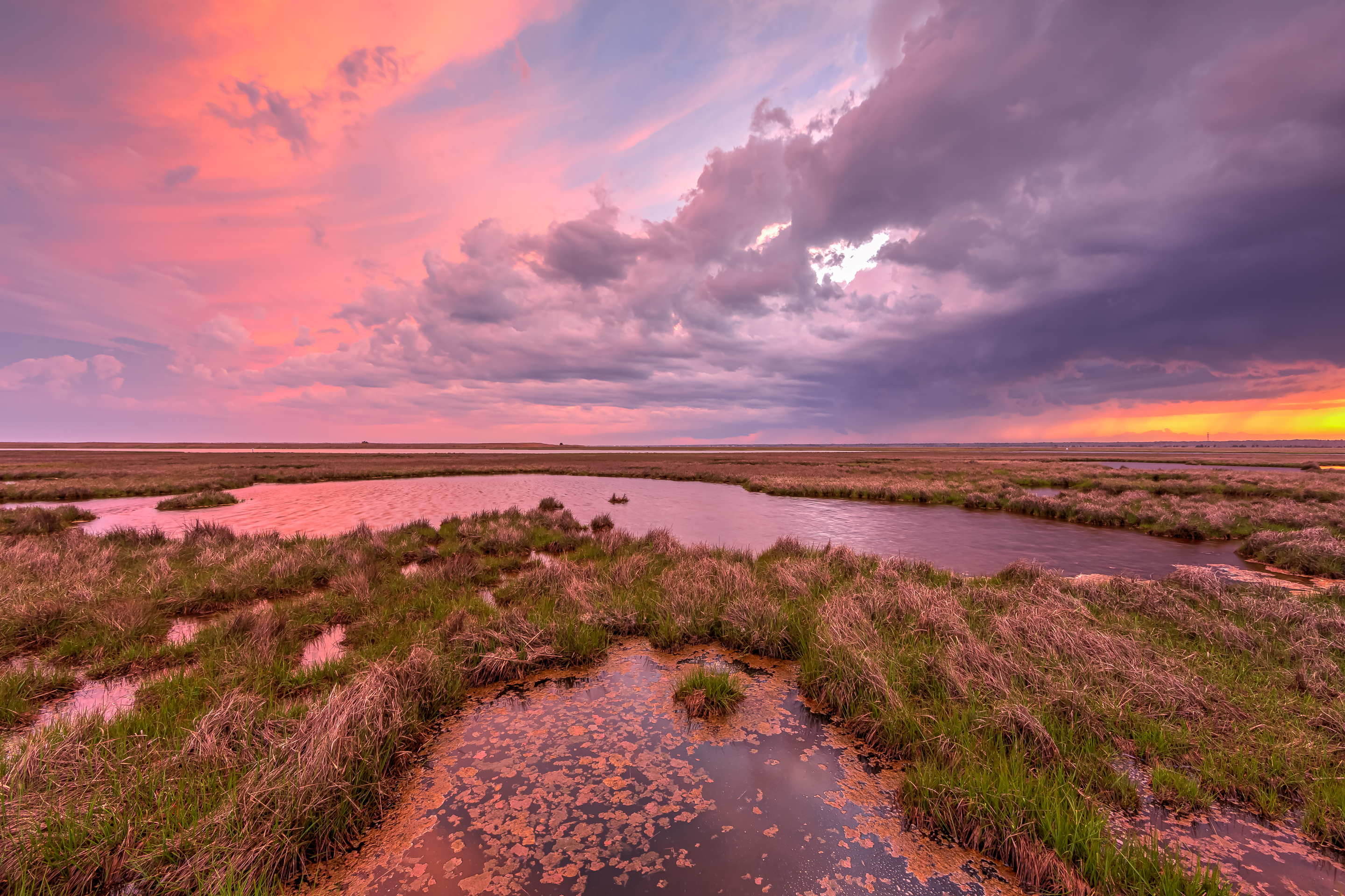 14mm sunset photograph over Cedar Run Dock Road salt marsh in mid May. The sky and clouds light up in pink pastel tones as storm clouds roll across the marsh from the western horizon.