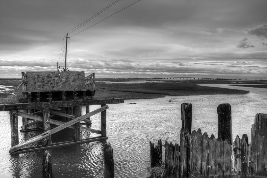 A weathered bridge structure beside water and marsh vegetation under an overcast sky.